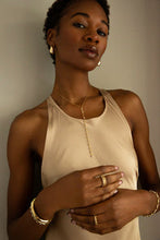 Load image into Gallery viewer, Against a neutral backdrop, a person with short hair wears a beige sleeveless top and brass jewelry. They are adorned with hoop earrings, layered necklaces, rings, and bracelets, including the Elongated Dome Earrings. Their relaxed pose shows them gently touching their necklace.
