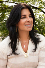 Load image into Gallery viewer, A woman with long black hair smiles while looking sideways, wearing a cream sweater and layered gold necklaces, featuring the Ball Chain Charm Necklace. Blurred greenery suggests she&#39;s outdoors, with sunlight warmly illuminating her face.
