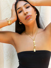 Load image into Gallery viewer, A woman, framed against a softly lit fabric backdrop, poses with her hands on her head. She&#39;s wearing gold hoop earrings, multiple bracelets, and a Lariat Toggle Charm Necklace. Her strapless black top beautifully contrasts with her long dark hair cascading freely.
