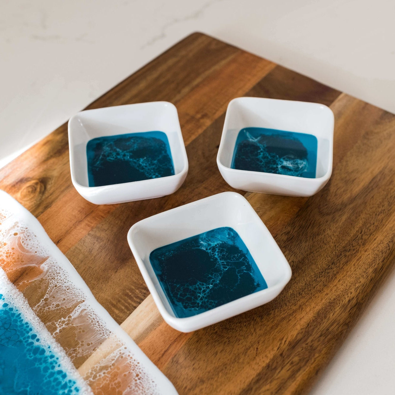 The Resin Accented Ceramic Dipping Bowls feature three square ceramic bowls filled with deep blue liquid and swirling patterns, set on a wooden board with ocean wave resin accents in blue, brown, and foam-like white resembling a seashore.