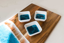 Load image into Gallery viewer, The Resin Accented Ceramic Dipping Bowls feature three square ceramic bowls filled with deep blue liquid and swirling patterns, set on a wooden board with ocean wave resin accents in blue, brown, and foam-like white resembling a seashore.
