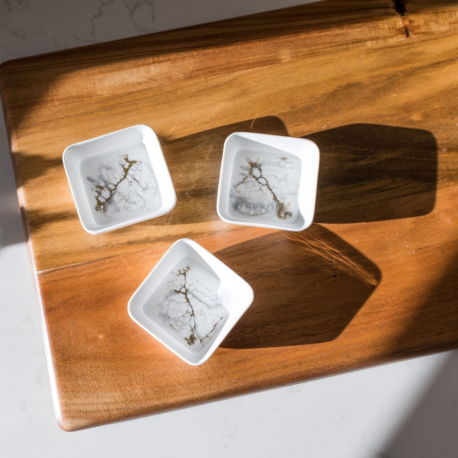 Three Resin Accented Ceramic Dipping Bowls with intricate floral patterns sit on a wooden cutting board on white marble. Sunlight casts long shadows, enhancing the scene's serenity and minimalist aesthetic with orderly patterns.