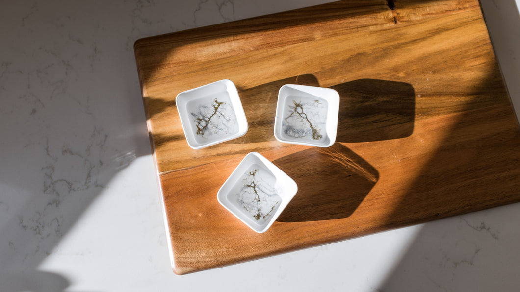 Three Resin Accented Ceramic Dipping Bowls with intricate floral patterns sit on a wooden cutting board on white marble. Sunlight casts long shadows, enhancing the scene's serenity and minimalist aesthetic with orderly patterns.