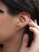 Load image into Gallery viewer, Close-up of a person&#39;s ear adorned with the Droplet Cross Studs, part of an elegant earring stack. The dark hair is tucked behind the ear, and they gently touch the back of the ear with their fingers. The skin is smooth, and the earrings subtly catch the light, highlighting their simplicity and elegance.
