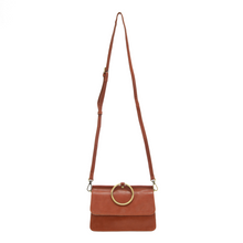 Load image into Gallery viewer, Introducing the Aria Ring Bag - Fall/Winter, a sophisticated brown vegan leather crossbody bag. It features a long adjustable strap secured with gold hardware on each side, complemented by a prominent gold ring on the front flap. Its rectangular silhouette and sleek design exude simplicity and elegance.
