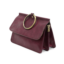 Load image into Gallery viewer, The Aria Ring Bag - Fall/Winter is a chic bracelet bag in burgundy, crafted from smooth vegan leather. It showcases a prominent large gold ring detail on the front flap. The bag includes a zippered side compartment and flaunts a structured, rectangular shape with a slightly glossy finish, elegantly positioned at an angle on the white background.
