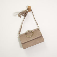 Load image into Gallery viewer, The Aria Ring Bag - Fall/Winter in taupe vegan leather is displayed hanging from a wooden peg against a simple white backdrop. This bag, echoing the style of a bracelet bag, boasts a prominent gold ring on its front flap and includes an adjustable shoulder strap, which is tied near the peg. Its design is both minimalist and modern.
