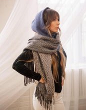 Load image into Gallery viewer, Wearing a black top and white pants, a woman places her hands on her hips, elegantly styled with the Cozy Scarves&#39; gray fringed design draped around her neck. She complements the look with a blue knit head covering. The warm lighting enhances the ambiance of the softly lit room with sheer curtains, while her long, wavy hair cascades gently over her shoulders.
