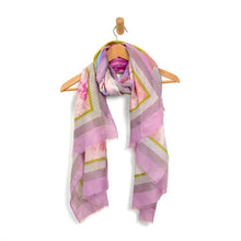 Load image into Gallery viewer, A Lightweight Patterned Scarf, adorned with geometric patterns and floral motifs in a vibrant array of colors, is elegantly draped on a wooden hanger. Dominated by purple hues, it showcases pink, green, and white accents and has fringed edges. The scarf is styled in a loose knot, symmetrically hanging down on both sides.
