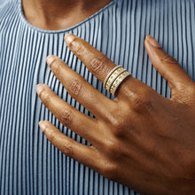 Load image into Gallery viewer, A hand with dark skin rests on a pleated blue fabric, adorned with the Two Tone 5-Stack Ring. This stunning piece features three 14K gold bands set with small, sparkling cubic zirconia stones that enhance the brilliance of the jewelry, offering a striking contrast to both the smoothness of the skin and the delicate texture of the fabric.
