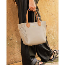 Load image into Gallery viewer, A person dressed in black pants and patterned sandals walks by a tan wall, carrying the fashionable Dublin Tote Bag made from vegan leather. The tote features brown handles, perforated side details, a small gold logo near the top, and a trendy ring accent on the side.
