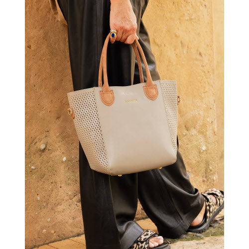 A person dressed in black pants and patterned sandals walks by a tan wall, carrying the fashionable Dublin Tote Bag made from vegan leather. The tote features brown handles, perforated side details, a small gold logo near the top, and a trendy ring accent on the side.