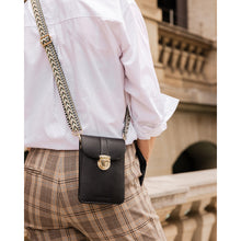 Load image into Gallery viewer, A person wearing a white shirt and plaid pants is seen from behind, carrying the Fontaine Phone Bag, a black leather crossbody with an adjustable patterned strap. They are standing near a stone railing with an ornate building in the background.
