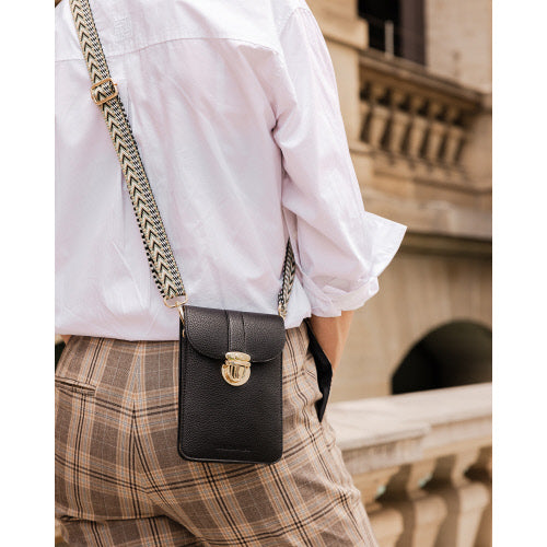 A person wearing a white shirt and plaid pants is seen from behind, carrying the Fontaine Phone Bag, a black leather crossbody with an adjustable patterned strap. They are standing near a stone railing with an ornate building in the background.