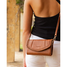 Load image into Gallery viewer, A woman dressed in a black sleeveless top and white pants is seen carrying the Harlow Crossbody Bag, made from vegan leather. The bag features intricate braided detailing on the flap and includes an adjustable crossbody strap for added convenience. The scene is set outdoors, with blurred pillars and greenery in the background.
