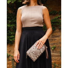 Load image into Gallery viewer, A woman in a sleeveless, ribbed taupe top and black skirt stands against an ivy-covered brick wall, holding the stylish Poppy Wristlet in vegan leather. Only her torso and lower face are visible, adding an air of mystery to her chic ensemble.
