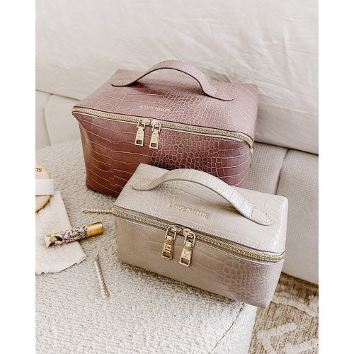 On a cream-colored surface, two Coordinating Toiletry and Makeup Cases with textured finishes are displayed. The top case, featuring a faux croc finish in burgundy, complements the elegant beige one below. Both cases include branded handles and gold-tone zippers. Accompanying them are a tube of lipstick and a small glass perfume bottle, enhancing the scene's elegance.