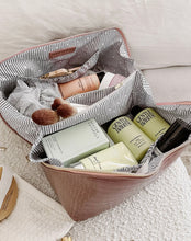 Load image into Gallery viewer, A brown leather Coordinating Toiletry and Makeup Case with a faux croc finish and striped interior is open, showcasing neatly arranged skincare and beauty items. Among them are bottles labeled &quot;Gentle Habits&quot; and a pink cylindrical container, alongside a makeup brush and a white mesh pouch. The case sits on a cream-colored textured surface.
