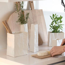 Load image into Gallery viewer, A kitchen counter features a person slicing cheese on a small wooden board, while Porcelain “Paper Bag” Vases resembling crumpled bags hold herbs and utensils. In the background, a wooden cutting board is present. Sunlight floods the area, creating a cozy and clean atmosphere.
