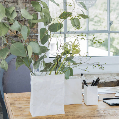 In front of a large window, there is a wooden table with a Porcelain “Paper Bag” Vase, designed to resemble a crumpled white paper bag, holding green leafy branches. Alongside it, another porcelain vase holds pens. A partially visible grey chair sits nearby, backed by a stone wall as sunlight filters through the window.