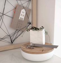 Load image into Gallery viewer, A minimalistic scene showcases a Porcelain “Paper Bag” Vase with cascading green vines resting on a wooden and white round container. A keychain bearing the label &quot;OPTIMIST&quot; and keys are placed on top. In the background, a birthday card adorned with a house design is clipped to a string art frame.
