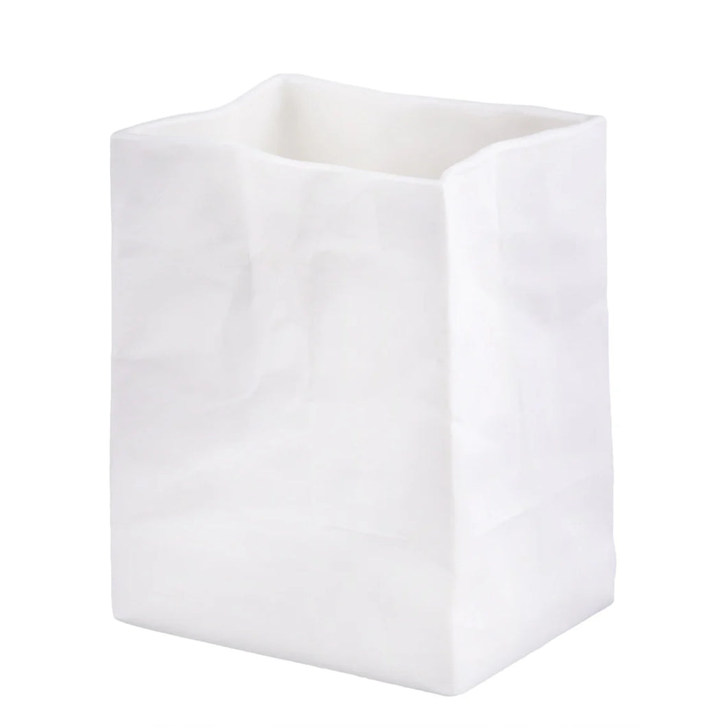 The Porcelain “Paper Bag” Vase is a white, bag-shaped vessel that stands upright, exuding an artisanal touch. Its surface is adorned with gentle creases and folds that mimic a real paper bag. This handcrafted porcelain piece features a rectangular base and a slightly uneven open top, making it a distinctive vase for any setting.