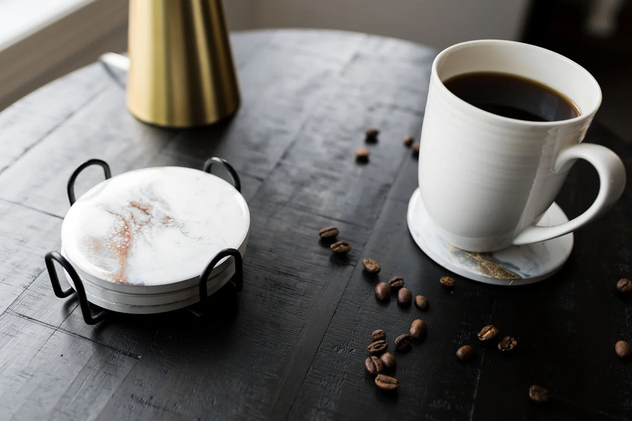 A white ceramic mug filled with black coffee sits on a coaster atop a dark wooden table. Coffee beans are scattered around, alongside the Ceramic Resin Coaster Set, featuring white and marbled stone coasters with non-slip cork backing nestled in a black holder. A gold-colored object is blurred in the background near a window.