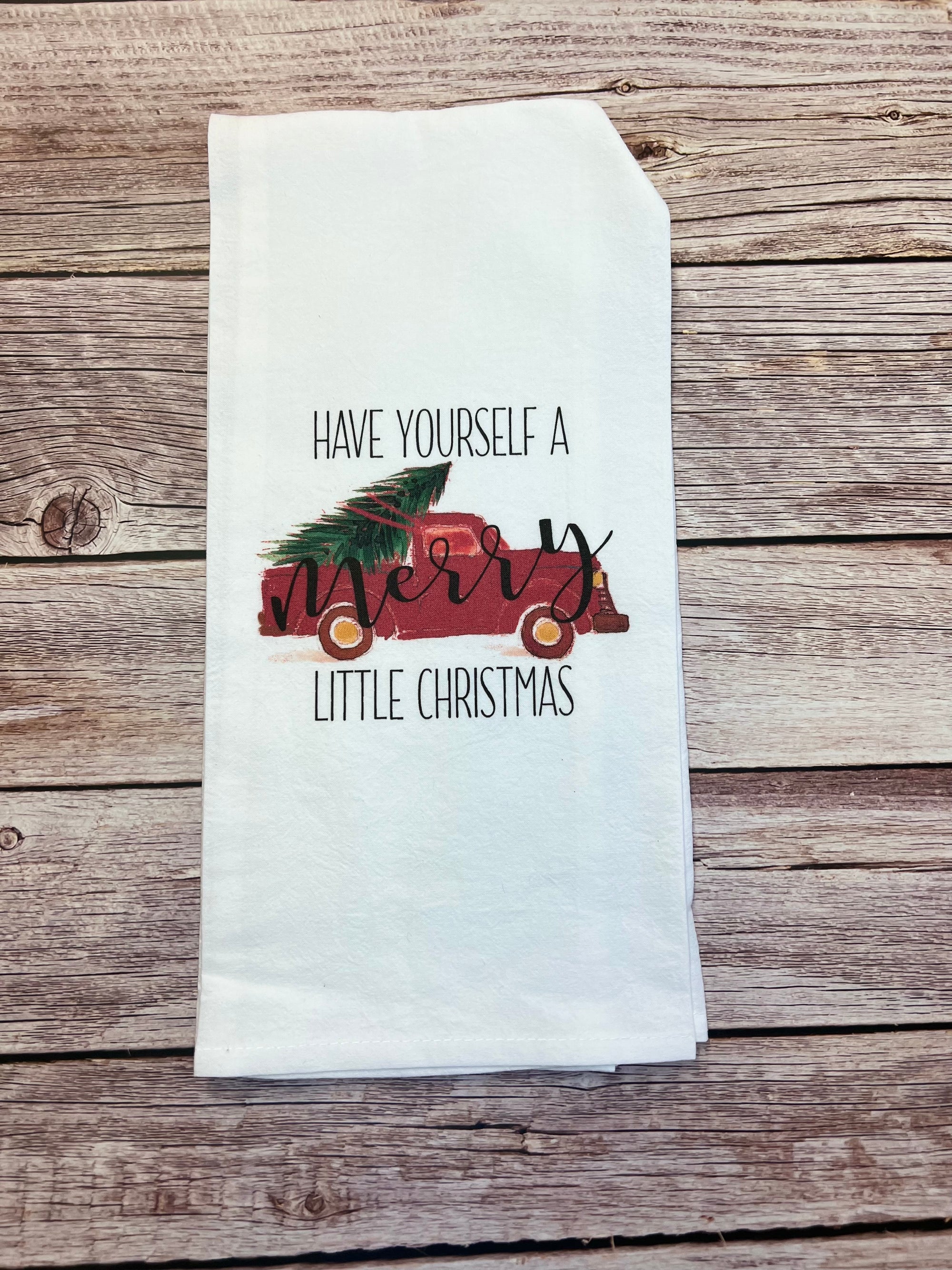 A tea towel from our Winter/Christmas collection showcases the phrase &quot;Have Yourself a Merry Little Christmas&quot; in black and red. The word &quot;Merry&quot; is beautifully decorated with green pine branches and red ornaments. This handcrafted item is displayed gracefully on a rustic wooden surface.