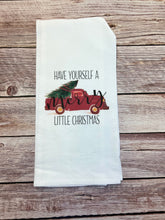 Load image into Gallery viewer, A tea towel from our Winter/Christmas collection showcases the phrase &quot;Have Yourself a Merry Little Christmas&quot; in black and red. The word &quot;Merry&quot; is beautifully decorated with green pine branches and red ornaments. This handcrafted item is displayed gracefully on a rustic wooden surface.
