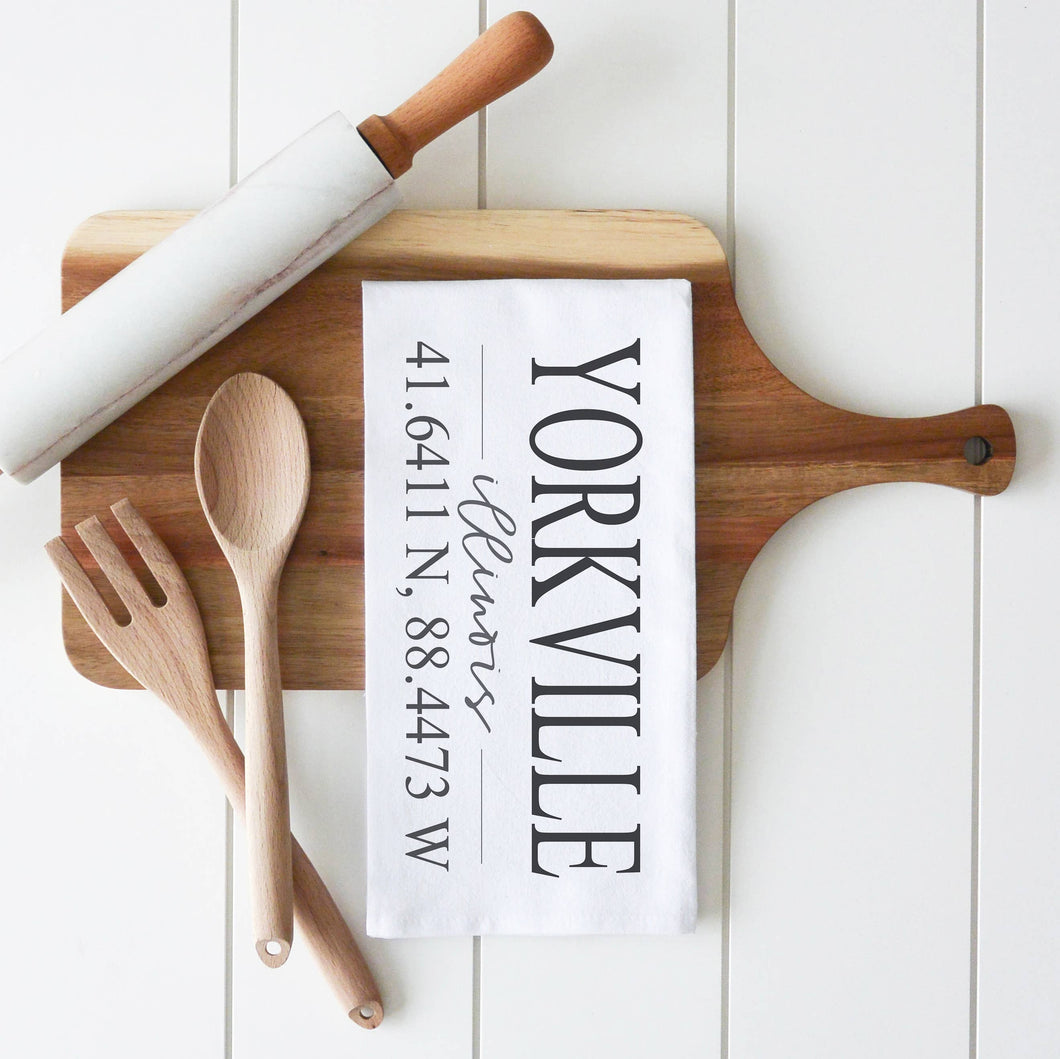 A wooden cutting board with a handle features a folded white tea towel from the 