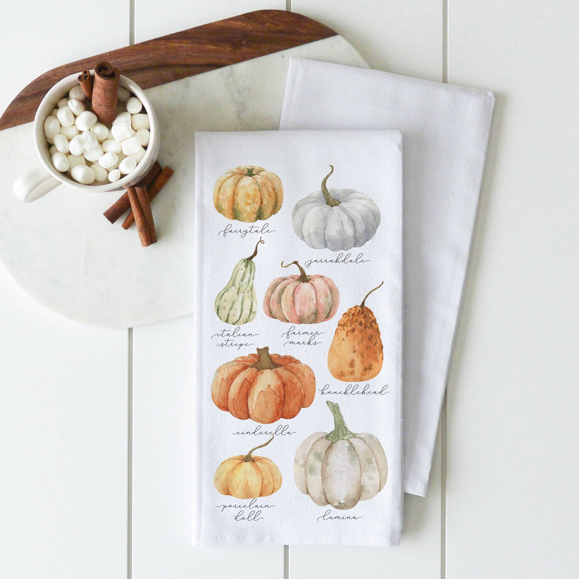 A Tea Towel from the Kitchen Collection's Fall/Thanksgiving series, adorned with watercolor illustrations of pumpkins such as "Musquee de Provence" and "Knucklehead," complements a white mug filled with marshmallows and cinnamon sticks, elegantly arranged on a white wooden surface. Crafted from high-quality materials, it exudes charm.