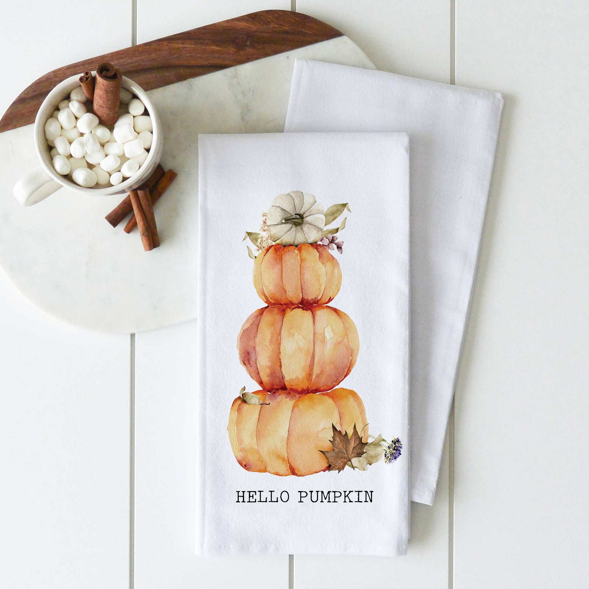 Part of our Tea Towels - Fall/Thanksgiving collection, this handcrafted white kitchen towel displays a charming watercolor illustration of three stacked pumpkins crowned with a small white pumpkin. Adorned with fall leaves and lavender sprigs, it features the cheerful message "HELLO PUMPKIN." Accompanying the towel is a cup brimming with marshmallows and cinnamon sticks.