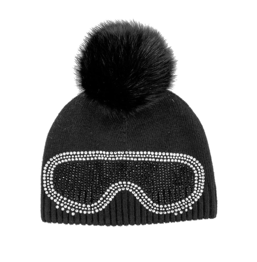 The Knit Beanie with Crystal Ski Goggles & Fox Fur Pom-Pom showcases a striking black design, highlighted by a large fox fur pom-pom on top. The front is adorned with sparkling rhinestone embellishments in the shape of ski goggles, offering both charm and fun. A ribbed texture along the bottom edge guarantees a secure and comfortable fit.