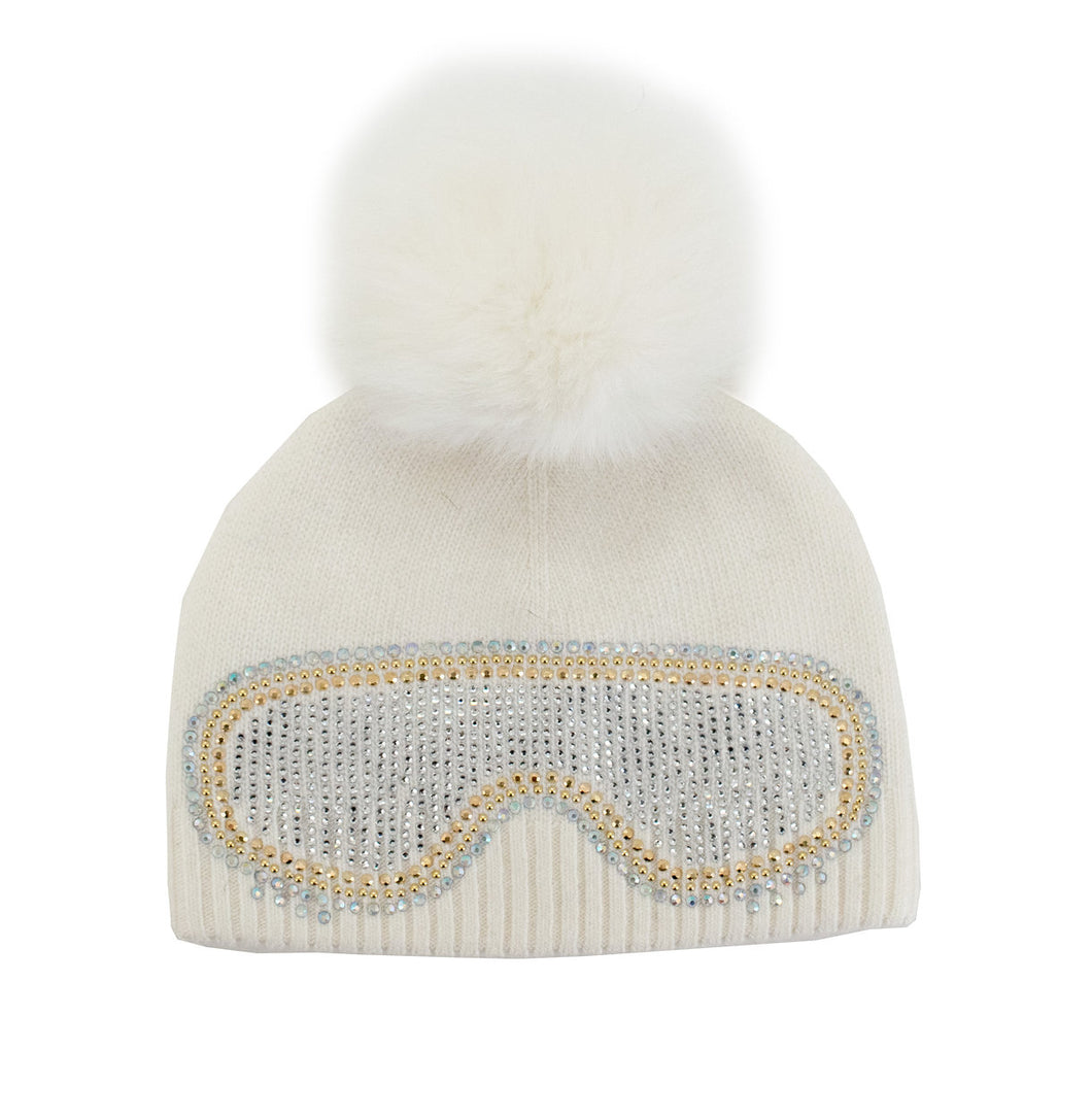 A sophisticated white knit beanie, topped with a luxurious fox fur pom-pom, features an eye-catching ski goggles design on the front, embellished with dazzling silver and gold rhinestones.