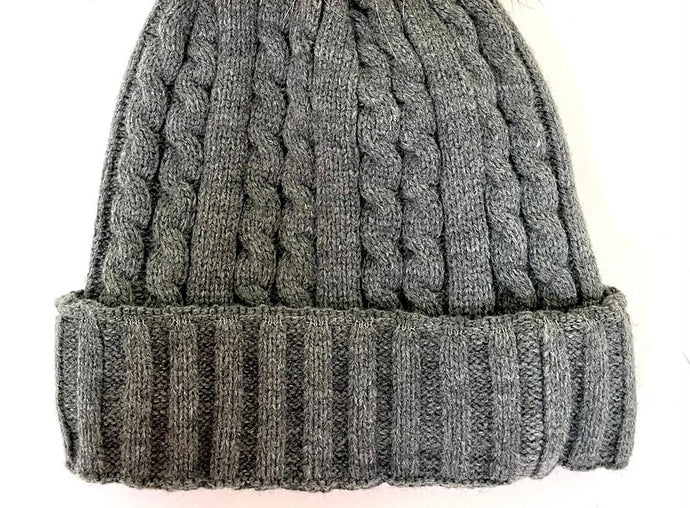 A gray Cable Knit Beanie with Fleece Lining & Fox Pom Pom features a ribbed fold-over brim and cozy fleece lining. Laid flat on a white surface, it beautifully showcases its intricate texture and design.