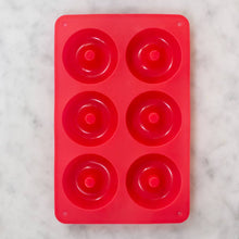 Load image into Gallery viewer, A red silicone mold from the Gingerbread Doughnut Baking Mix &amp; Mold Set, featuring six circular cavities, rests on a white marble surface. Each cavity includes a raised center for creating the classic doughnut hole, while two small round holes at each corner facilitate easy handling or hanging. It&#39;s perfect for making delicious Gingerbread Doughnuts.
