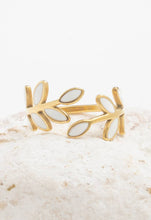 Load image into Gallery viewer, The Seeds of Hope Ring, a stunning open design piece with 14K gold plating, features two rows of white enamel leaves elegantly framing each side. This adjustable ring is beautifully displayed on a textured, light-colored stone set against a plain white background.
