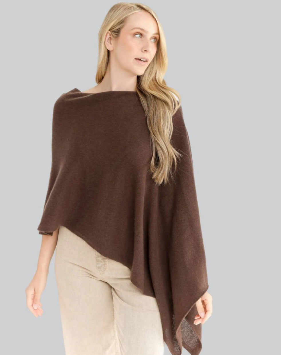 A woman with long blonde hair is wearing the Cashmere Poncho in brown, styled asymmetrically over beige pants. She stands against a light gray background, gazing to the side with a relaxed expression. The one-size dress topper drapes elegantly, highlighting its soft texture.