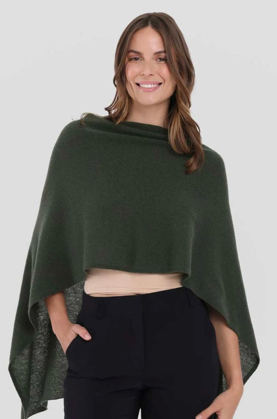 A woman with long brown hair smiles while wearing the dark green Cashmere Poncho over a beige top. She pairs it with black pants and casually places one hand in her pocket. The plain white background accentuates the elegance of her versatile, one-size outfit.
