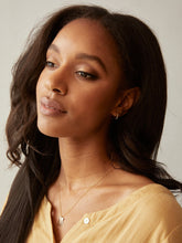 Load image into Gallery viewer, A woman with long, dark hair gazes to the side against a neutral background. She is wearing a light yellow blouse, 14k gold-filled Celine Huggie Hoops, and a delicate necklace featuring a rectangular pendant. Her makeup is understated, highlighting her eyes, and she has a calm, thoughtful expression.
