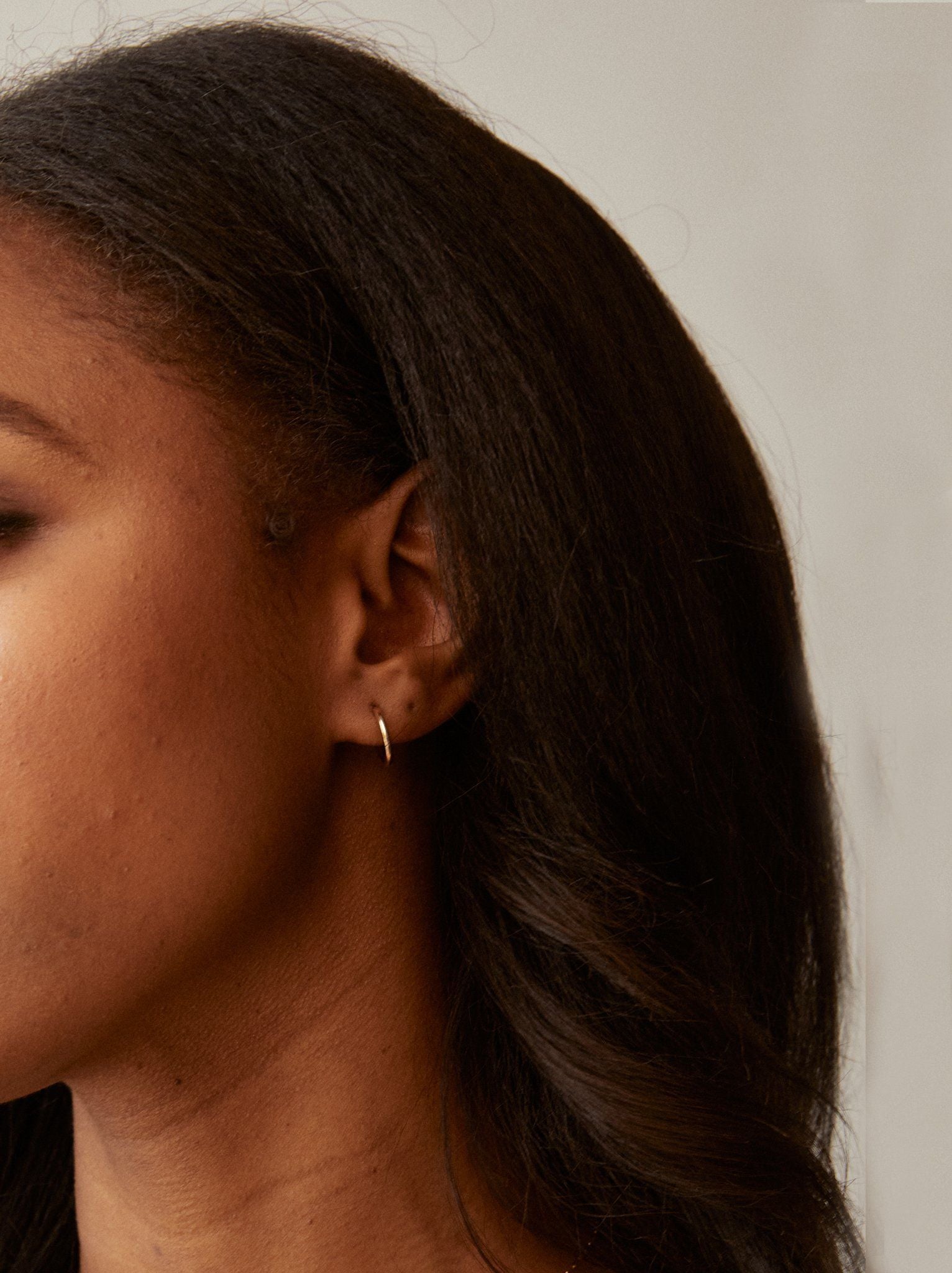 Close-up of the left side of a woman's face with long, dark hair. She is adorned with a Celine Huggie Hoop crafted from 14k gold-filled material. The background is a neutral, soft-focus surface that accentuates her ear, the earring from an ethical fashion brand, and her cascading hair.