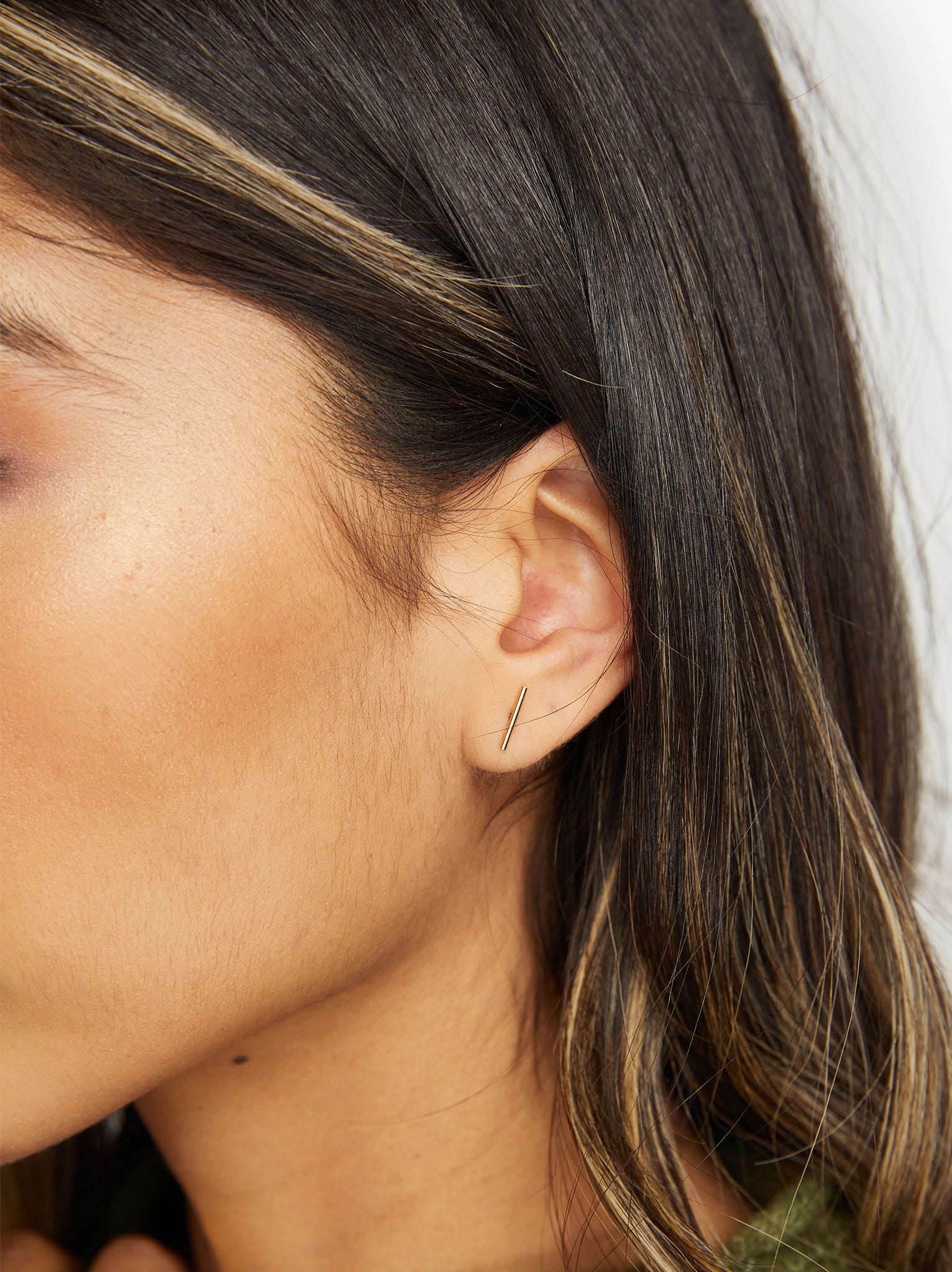 A close-up showcases a person's left ear featuring the minimalist Stick Studs, which reflect ethical fashion values. These thin gold bar earrings belong to a handmade jewelry collection. Straight dark hair with light highlights cascades past the ear and against tan skin, emphasizing the blurred background to highlight the earring.