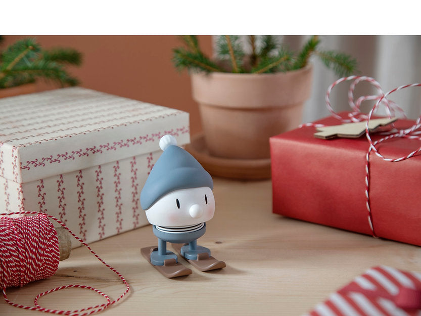 The Hoptimist Skiing Bumble, a small toy figure crafted from ABS plastic and sporting a blue hat, stands on skis among wrapped gifts. Nearby, red and white twine adds to the charm, while potted plants in the background enhance the festive atmosphere of the winter landscape.