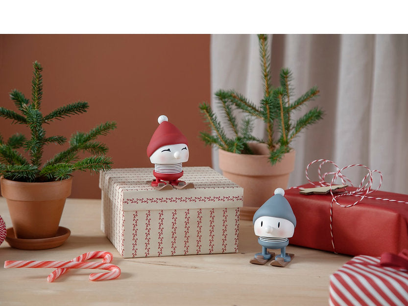 The image features two Hoptimist Skiing Bumble figurines made of small ABS plastic, each wearing red and blue hats, perched on gift boxes wrapped in red and white paper. Surrounding them are potted evergreen plants, candy canes, and string decorations that evoke a winter landscape ambiance. In the background, a red wall pairs with a beige curtain for contrast.