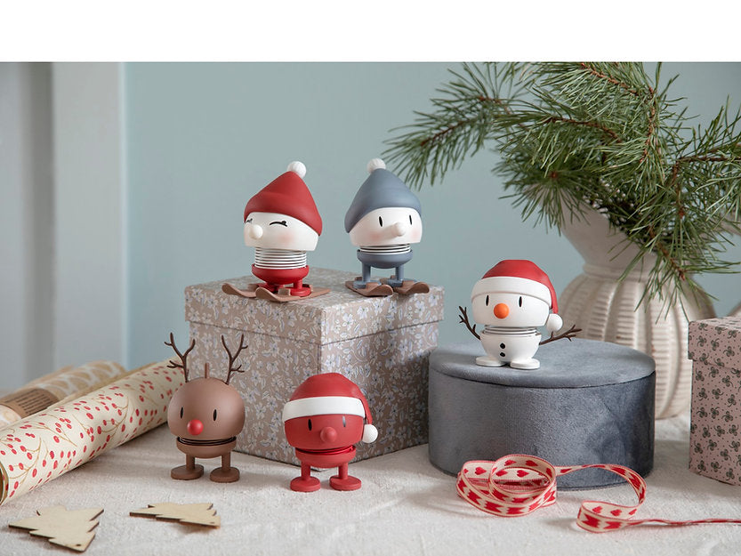 A festive scene showcasing the Hoptimist Skiing Bumble collection: two with red hats perched on a patterned box, one adorned in a gray hat seated atop a silver box beside a small potted pine tree, creating a winter landscape. There's also a cheerful snowman figure and two figurines below, one sporting reindeer antlers. Rolls of ribbon and paper are scattered around them.