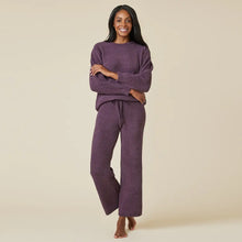 Load image into Gallery viewer, A person with long hair, barefoot against a plain backdrop, smiles while wearing the Marshmallow Lounge Set in purple. It features a knit sweater and elasticized drawstring pants. Their arms are crossed, highlighting the reverse seam shoulder stitching, and they appear relaxed.
