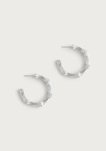 Load image into Gallery viewer, The Bamboo Medium Hoop Earrings - Silver by Anabel Aram are crafted from sterling silver plated brass, featuring a bamboo-like texture and segmented sections for a natural, wood-inspired look against a light, neutral background.
