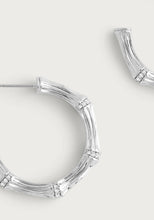 Load image into Gallery viewer, A close-up of the Bamboo Medium Hoop Earrings - Silver by Anabel Aram. Made from sterling silver plated brass, these earrings mimic bamboo segments with a textured finish and small sparkling stones at each joint, partially visible against a light background.
