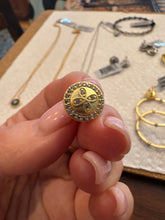 Load image into Gallery viewer, A person holds a round Clover Stud Earring with a four-petal flower design encrusted with small gems and bordered by cubic zirconia. In the background, a display showcases various necklaces and bracelets on a textured surface.
