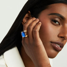 Load image into Gallery viewer, A person with long black hair and smooth brown skin gazes sideways, softly touching their face adorned with the Plaza Ring. They wear a white garment against a pale, solid backdrop.
